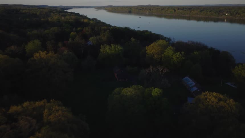 A serene aerial view glides over a lush forest toward a secluded house on the banks of a wide river at sunrise