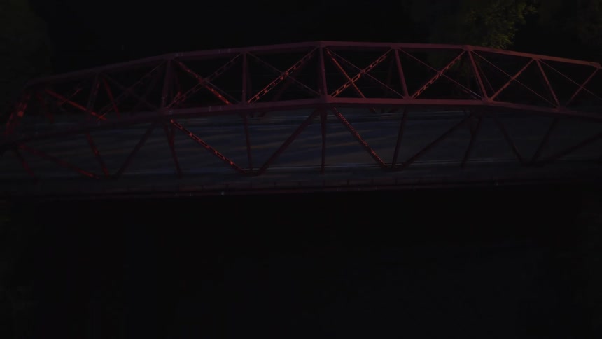 A beautiful aerial shot flies over a quaint small town at sunset, slowly pushing in and tilting down towards a classic red truss bridge over a calm river with a waterfall nearby