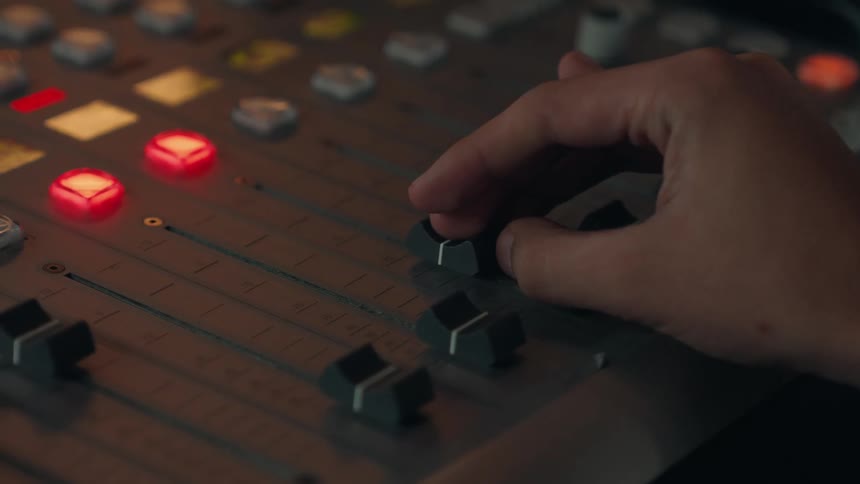 A sound producer's hand finely tunes the audio levels on a professional mixing console with glowing lights
