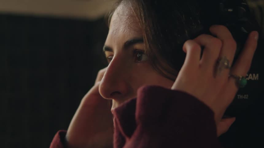 A close-up profile of a woman with dark hair putting on studio headphones in a moody, dimly lit setting