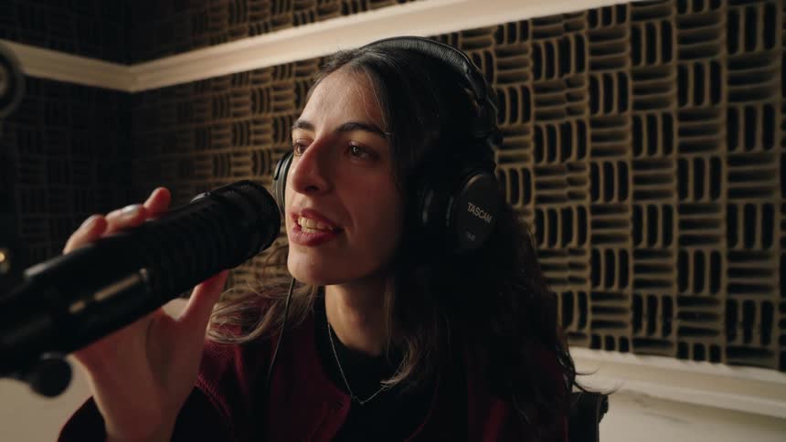 A young woman puts on her headphones and begins speaking into a microphone in a professional recording studio