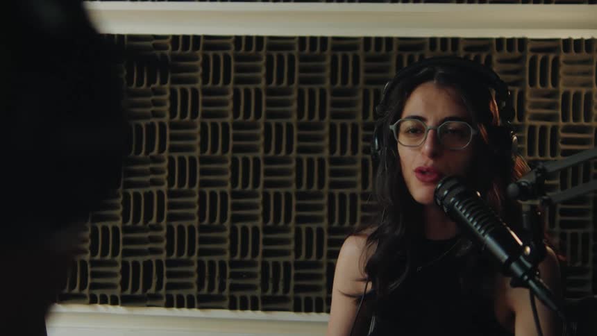A woman with glasses and headphones speaks into a microphone during a podcast recording session in a soundproof studio
