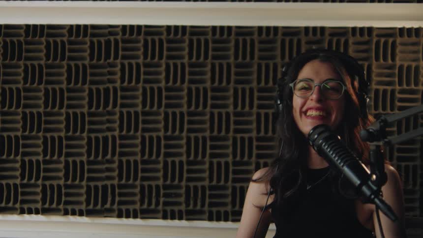 A joyful woman with glasses and headphones laughs heartily while recording a podcast in a soundproof studio