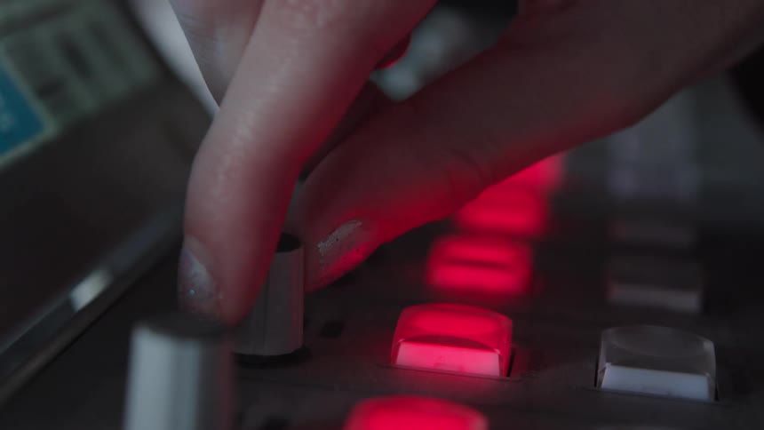 A musician's hand with sparkly nail polish presses glowing red buttons and turns a knob on an electronic music controller