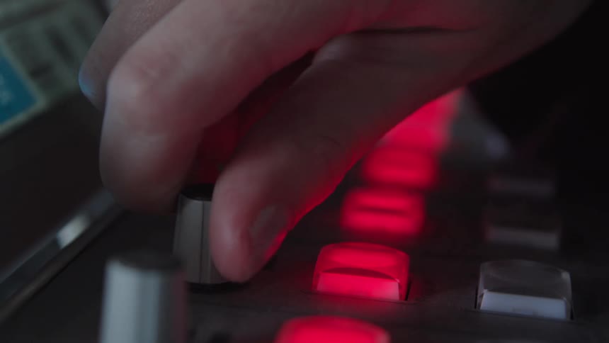 A close-up shot of a hand turning a knob on a music controller with glowing red buttons in a dimly lit room