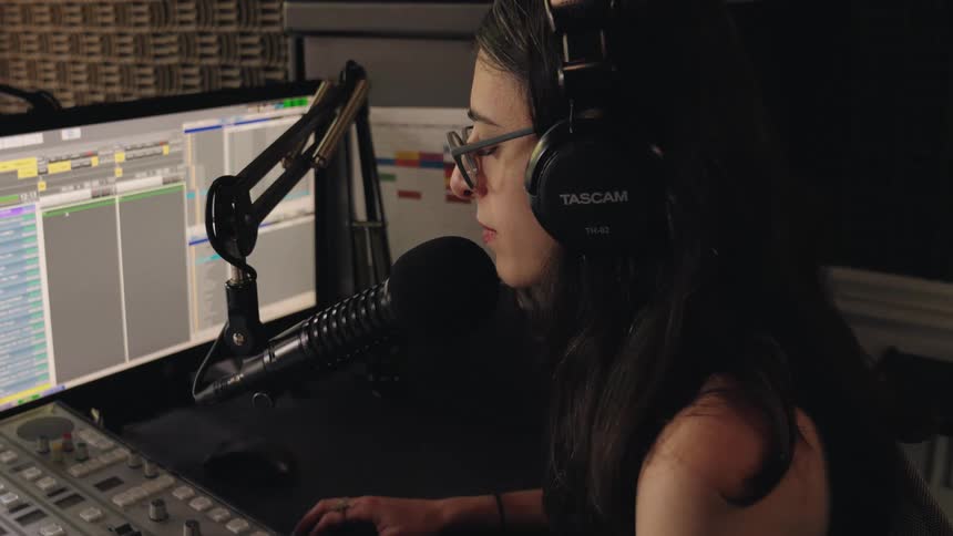 A young woman with glasses and headphones speaks into a microphone while working at a soundboard in a professional recording studio
