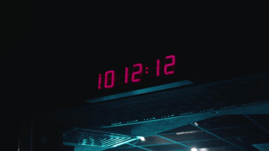 A pink digital clock counts the passing seconds in a dark, moody room as the camera slowly zooms in
