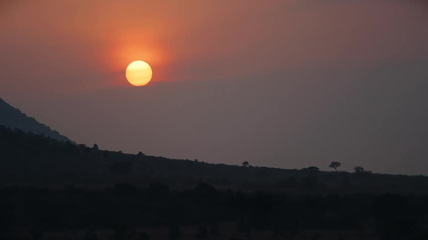 A vibrant orange sun hangs in the hazy sky as it sets behind the silhouetted hills of the African savanna