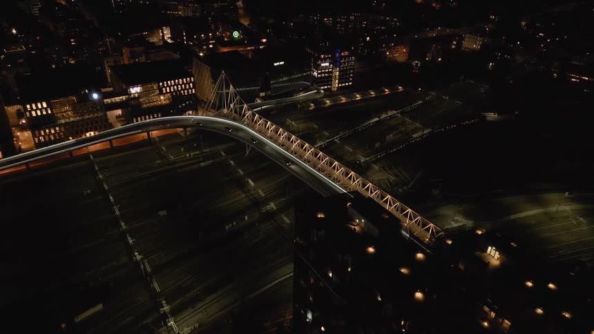 A striking aerial view shows a modern illuminated bridge with traffic crossing over a dark railway yard in a sprawling city at night