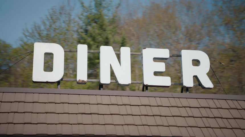 Tracking shot of a classic white roadside diner sign with trees and blue sky in the background