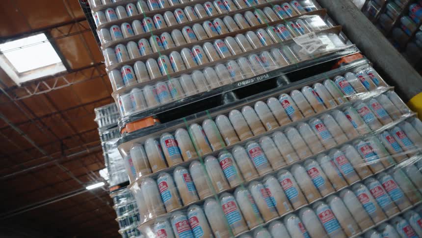 A slow pan reveals towering stacks of shrink-wrapped beer cans on pallets in a distribution warehouse