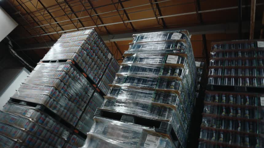 A slow, low-angle shot reveals massive stacks of shrink-wrapped canned beverages on pallets reaching towards the ceiling of a large distribution warehouse