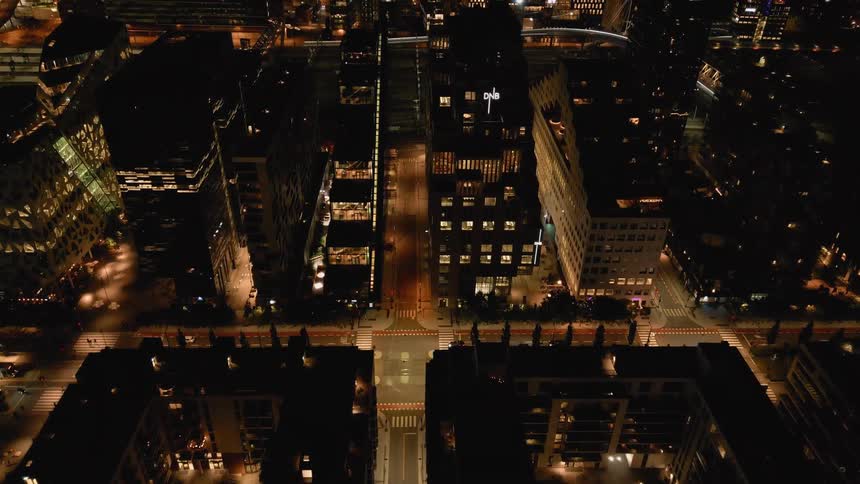 An aerial top-down view of a modern city street at night, slowly tilting up to reveal the illuminated urban landscape