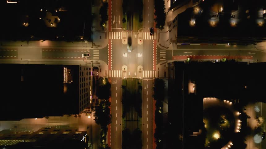 A cinematic top-down aerial shot slowly rises over a modern, symmetrical city street and intersection illuminated by warm lights at night