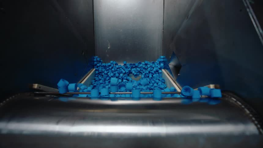 A torrent of blue plastic bottle caps falls onto a conveyor belt in a manufacturing plant, captured in slow motion
