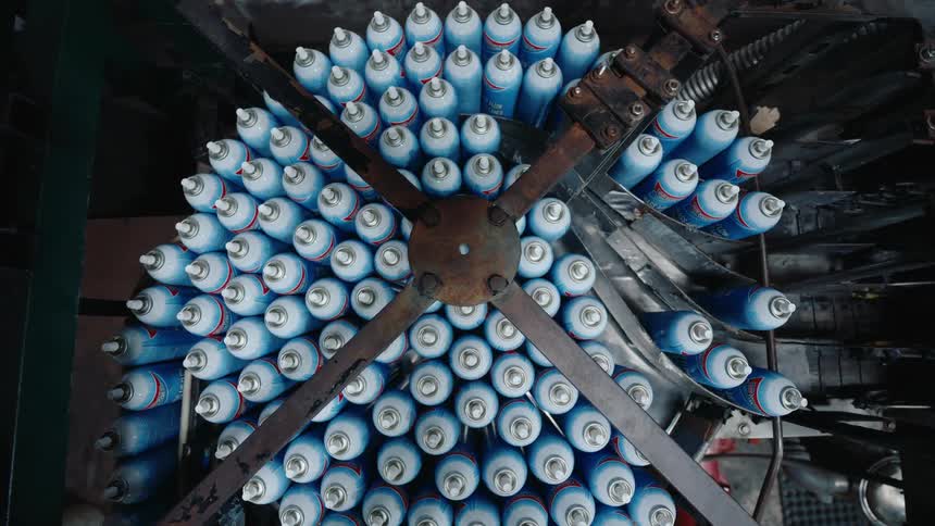 An industrial machine on an assembly line sorts hundreds of blue aerosol cans in a factory, seen from a top-down perspective