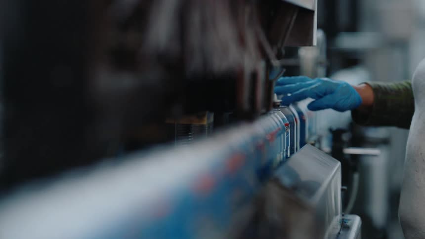 A worker's gloved hand guides aerosol cans along a fast-moving factory production line in slow motion