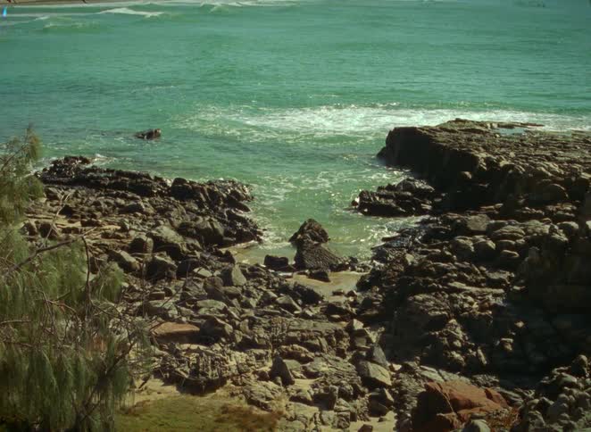 A vintage-style film shot of a serene rocky cove with turquoise ocean waves lapping at the shore