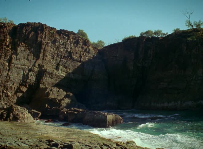 Slow-motion footage shows turquoise ocean waves crashing against the rugged rocks of a secluded sunlit cove