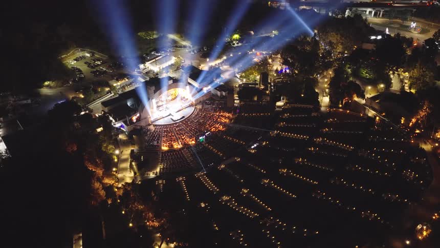 An expansive aerial shot captures a spectacular night concert at the Hollywood Bowl, with vibrant spotlights piercing the dark sky over a packed audience