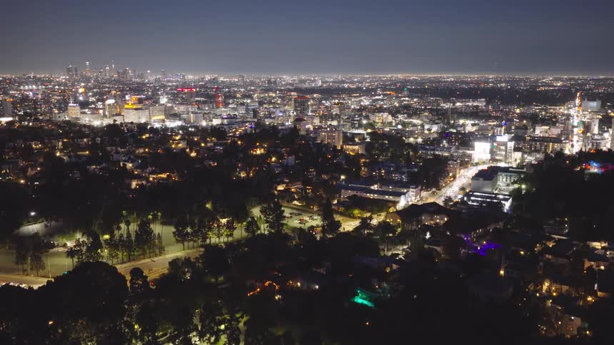 A slow aerial rise reveals the sprawling, brightly lit cityscape of Los Angeles at night, with the downtown skyline on the horizon