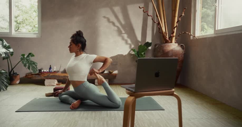 A woman participates in an online yoga class, holding a deep stretch pose on her mat in a calm, sunlit studio