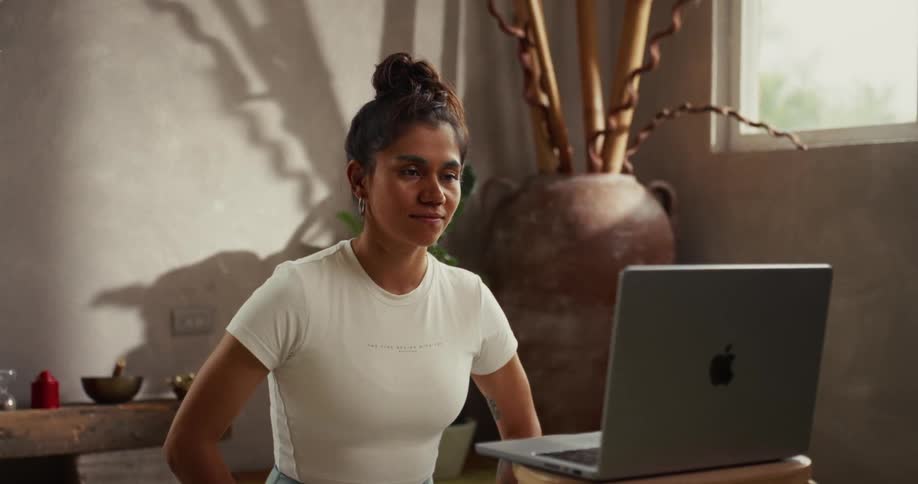A serene woman with her hair in a bun follows a guided meditation on her laptop in a sunlit room