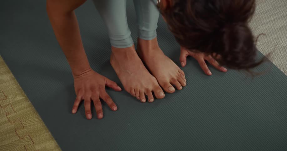 A yogi holds a standing forward fold pose, with a close-up on their bare feet and hands grounded on a yoga mat