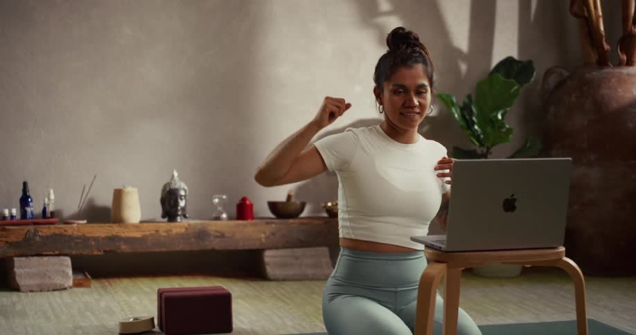 A smiling woman in a calm, zen-like studio finishes a stretch while following a virtual yoga class on her laptop
