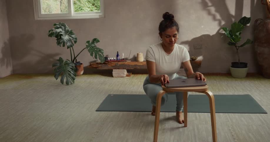 A barefoot woman in workout clothes walks in slow motion to set up her laptop on a stool before her online yoga class