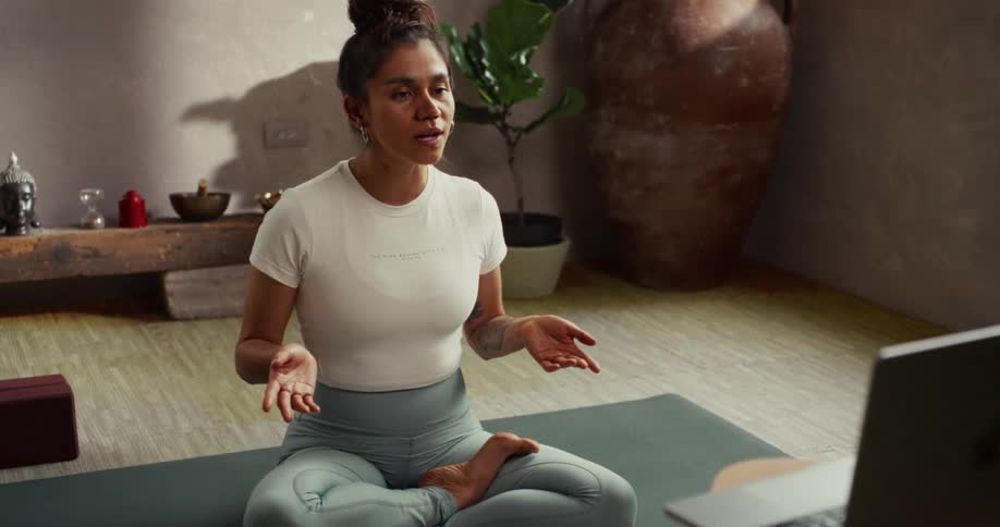 A female yoga instructor sits in the lotus position on her mat, teaching a virtual meditation and breathing class via her laptop in a tranquil studio