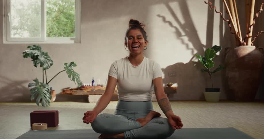 A smiling woman in a lotus position on a yoga mat begins to laugh joyfully in a sunlit studio
