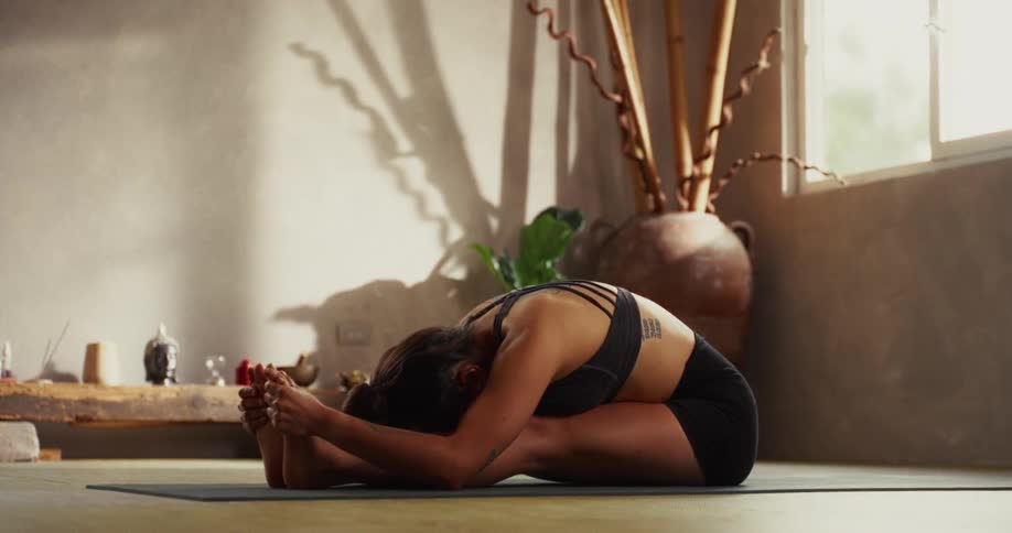 A woman practices a deep seated forward bend yoga pose in a tranquil, sunlit studio
