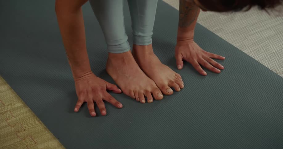 Slow motion close up of a woman's hands and feet as she practices a standing forward bend on a yoga mat