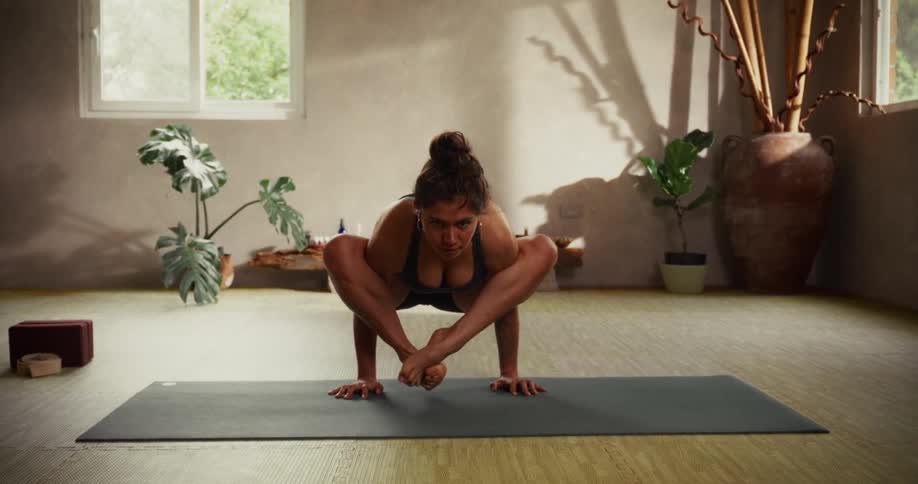 A strong and flexible woman with a back tattoo holds a difficult arm balance yoga pose in a serene studio