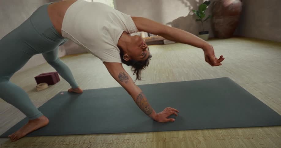 A tattooed woman gracefully transitions into a wild thing yoga pose with fluidity and control in a sunlit studio