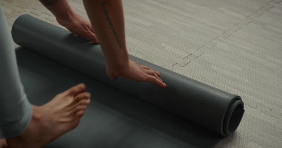 A person with tattooed arms carefully unrolls a grey yoga mat on the floor in slow motion