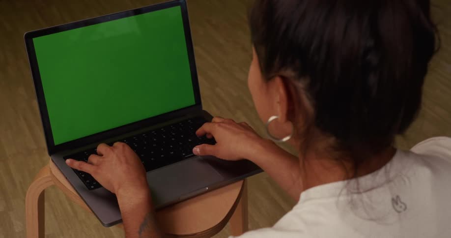 An over-the-shoulder shot of a woman typing on a laptop computer with a green chroma key screen