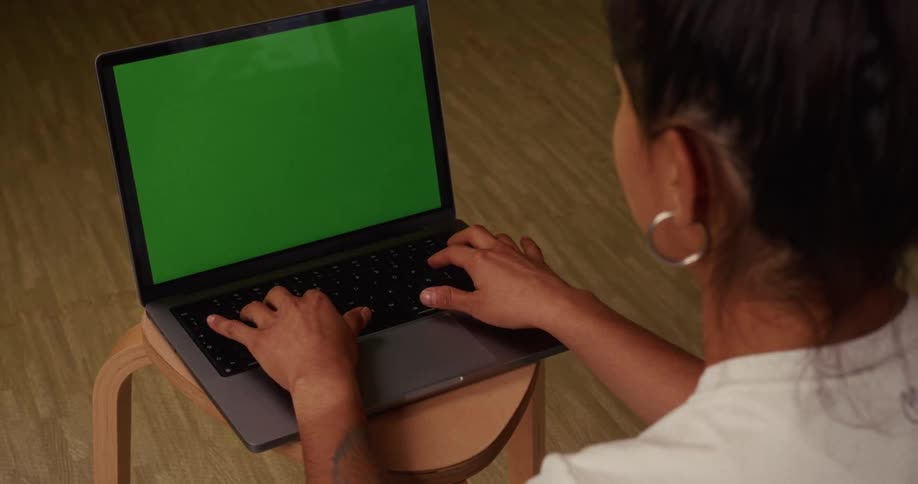 An over the shoulder view of a woman sitting on a stool and typing on a laptop with a green screen