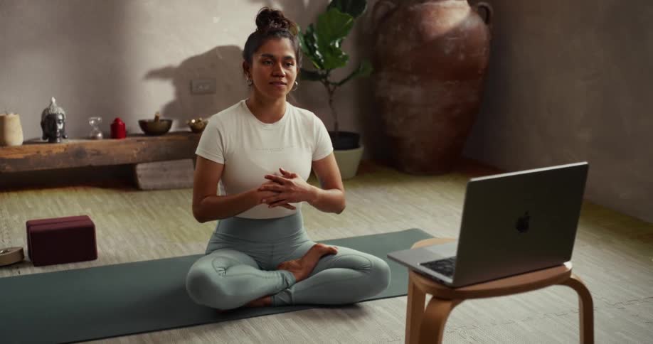 A calm yoga instructor sitting in the lotus position on a mat guides an online meditation class on her laptop in a serene studio