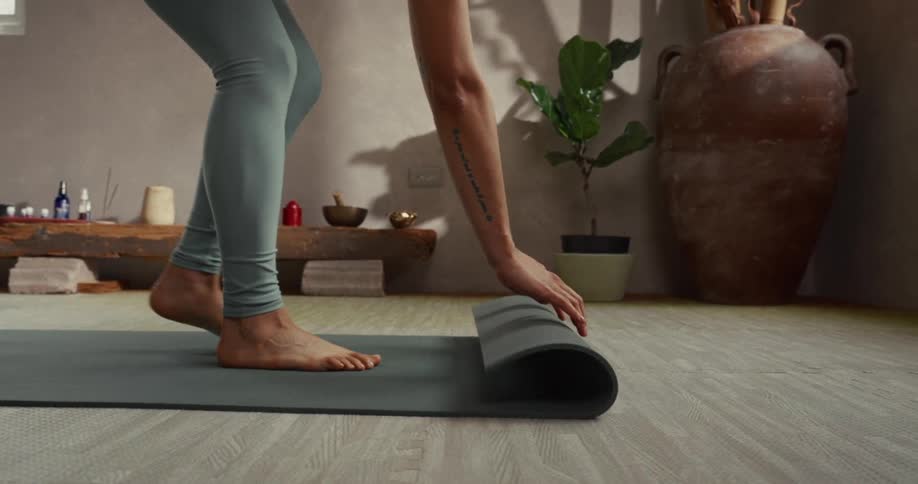 A woman in leggings slowly and gracefully rolls out her yoga mat on the wooden floor of a peaceful studio