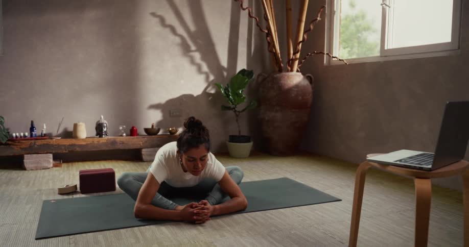 A woman peacefully follows an online yoga class on her laptop, folding forward into a stretch on her mat in a sunlit studio