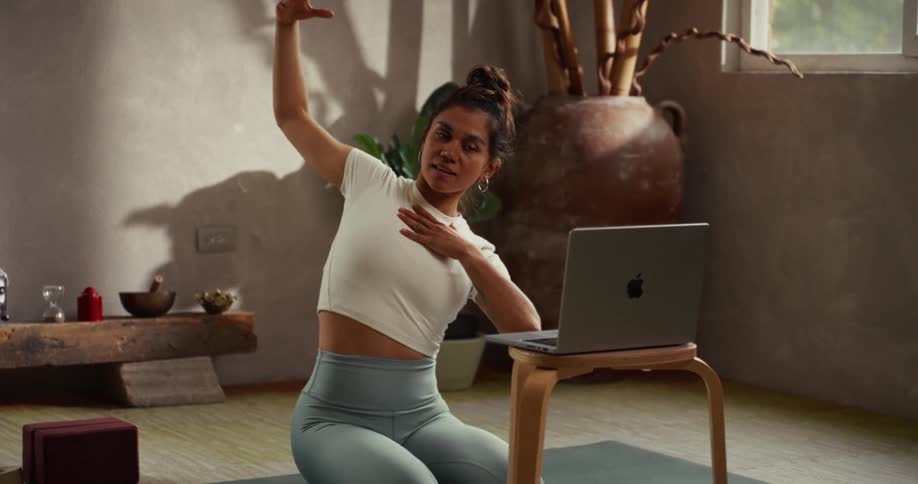 A woman in a tranquil, sunlit room gracefully performs yoga stretches in slow motion while following a virtual class on her laptop