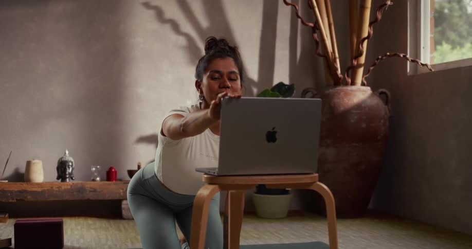 In a tranquil, sunlit studio, a smiling female yoga instructor waves goodbye to her virtual class before gently closing her laptop