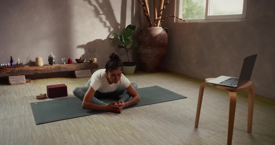 A calm woman practices a seated forward fold yoga pose on her mat while following instructions from an online class on her laptop