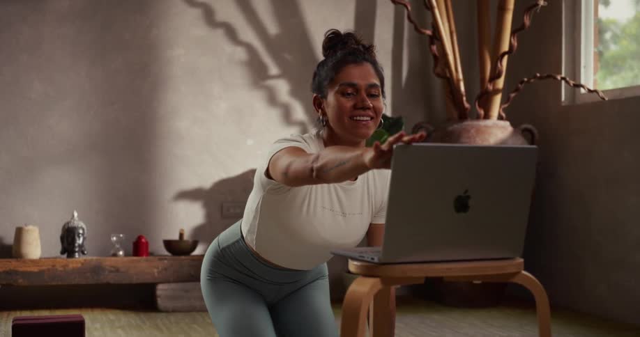 A smiling woman in a tranquil home studio finishes her online yoga class by waving goodbye and closing her laptop