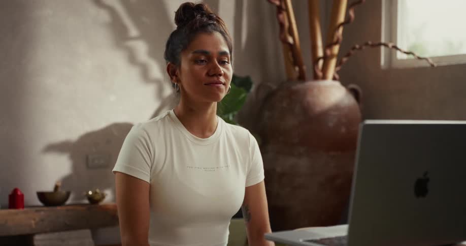 A smiling woman with her hair in a bun sits in a sunlit room and interacts with someone during a video call on her laptop