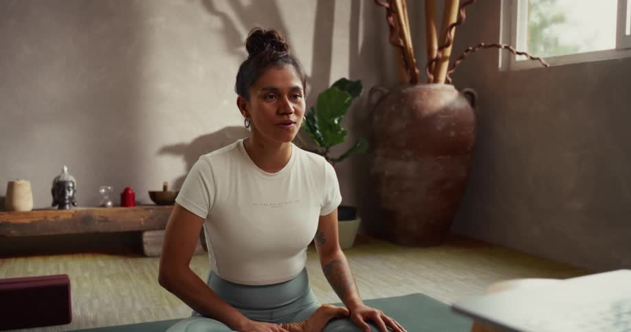 A serene yoga instructor sits on her mat in a lotus pose, leading an online class from her peaceful, sunlit studio