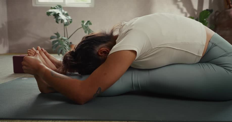 A flexible woman performs a deep seated forward bend yoga stretch on a mat in a serene, sunlit room