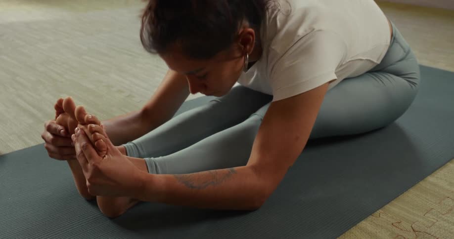 A woman slowly performs a seated forward bend, stretching her hamstrings on a yoga mat in a studio